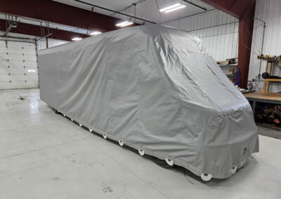 Large recreational vehicle or camper inside a warehouse fully covered in a custom-fitted light grey protective tarp.