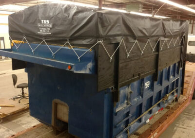 Large blue open-top container covered with a heavy black tarp secured by zig-zag shock cord lacing.