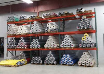 Multi-tier industrial warehouse rack stacked with various colored rolls of heavy-duty tarp fabric.