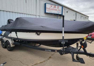 Profile view of a white boat hull wrapped tightly under a black marine canvas travel cover in front of a workshop.