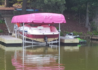 A lakeside speed boat sitting on a mechanical lift station under a striking, high-visibility bright pink custom canopy top cover.