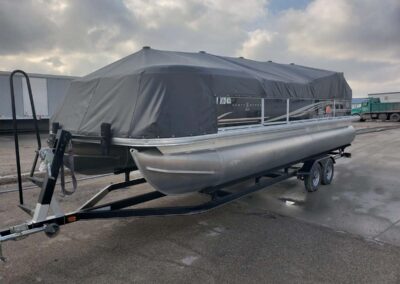 Dark gray travel cover fitted onto a pontoon boat on a transport trailer.