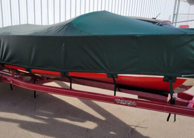 Side shot of a boat on a bright red trailer parked outdoors, utilizing a tightly secured forest green custom tie-down travel cover.
