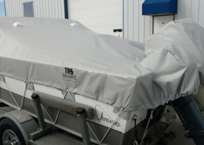 Side profile view of a boat on a trailer using a gray custom marine cover secured to the hull with a lashing hook system.