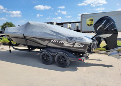 Side-profile view of a white boat on a dual-axle trailer covered by a gray ratchet-style travel cover extending over a large outboard motor.