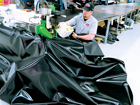 industrial-vinyl-welding-tarp-production A technician operating a commercial Miller Weldmaster rotary hot air vinyl welding machine, joining panels of heavy black industrial tarp vinyl in a clean manufacturing facility.