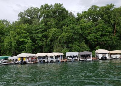 A scenic wide-angle view showing a long row of various custom boat lift canopy covers lined up along a forested lake shoreline.