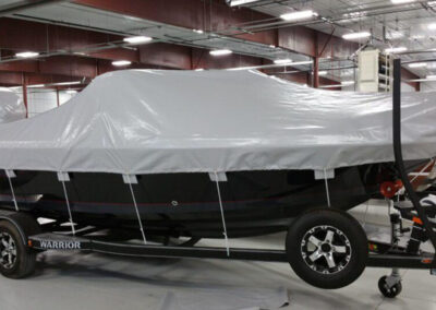Profile view of a large boat on a dual-axle roller trailer protected by a tightly tied-down light gray trailer-mount cover.