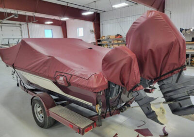 Side profile view of a boat on a trailer inside a workshop, protected under a custom maroon marine canvas ratchet cover.