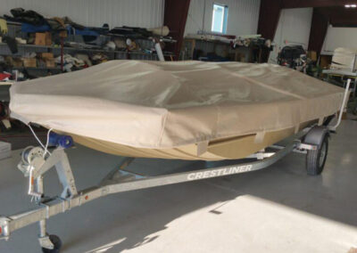 Angled view of a fiberglass boat on a single-axle trailer inside a workshop, protected under a tan tie-down storage cover.