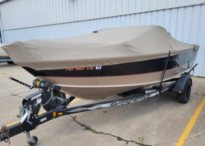 Side perspective view of a sport boat on a trailer protected by a dark tan custom travel cover inside a workshop garage.
