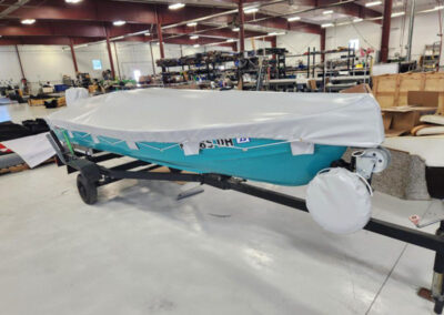Side profile of a boat with a distinct teal hull stripe, enclosed in a tailored light gray canvas storage cover with hull attachments.